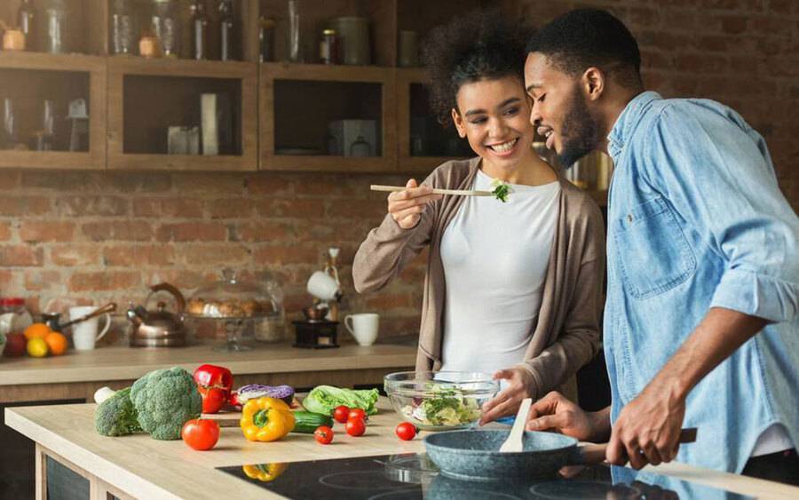 A young woman holds up a wooden spoon with food to her partner, representing healthy lifestyle changes.