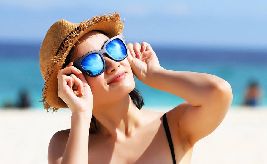 A lady sitting at the beach looks up toward the sun with a hat and sunglasses to protect her eyes from damage from the sun's rays.