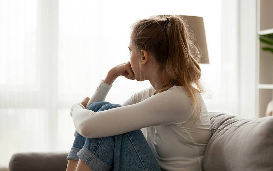 A teen sitting on the couch looking out the window looking depressed.
