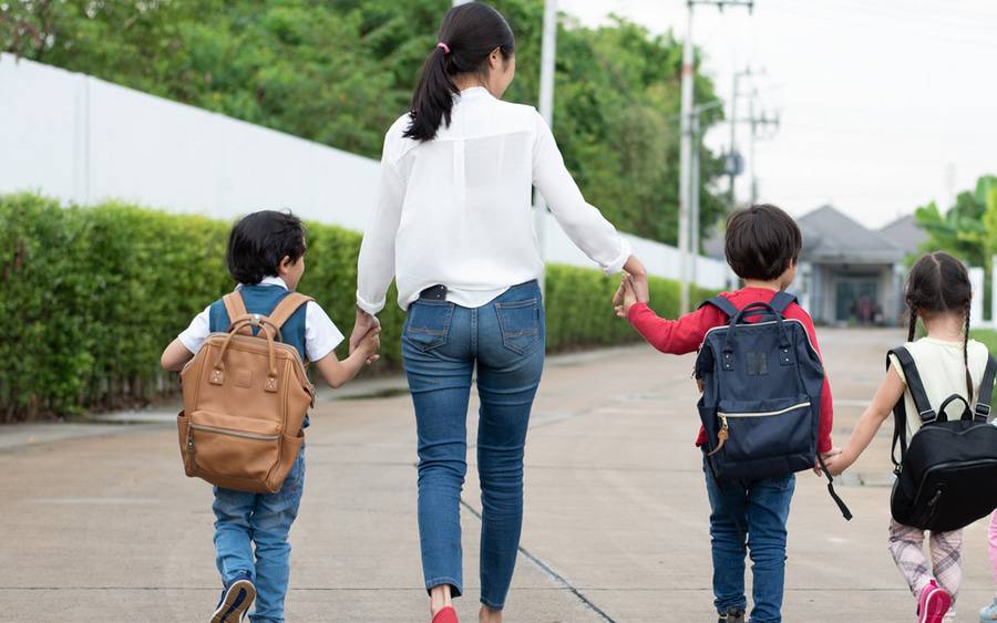 A woman walks hand-in-hand with three young children wearing backpacks.