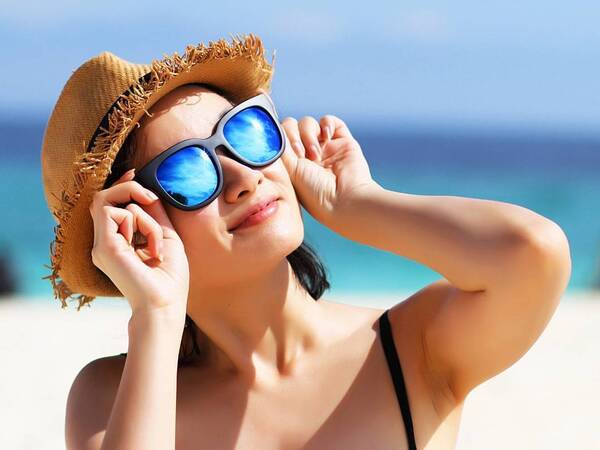 A lady sitting at the beach looks up toward the sun with a hat and sunglasses to protect her eyes from damage from the sun's rays.