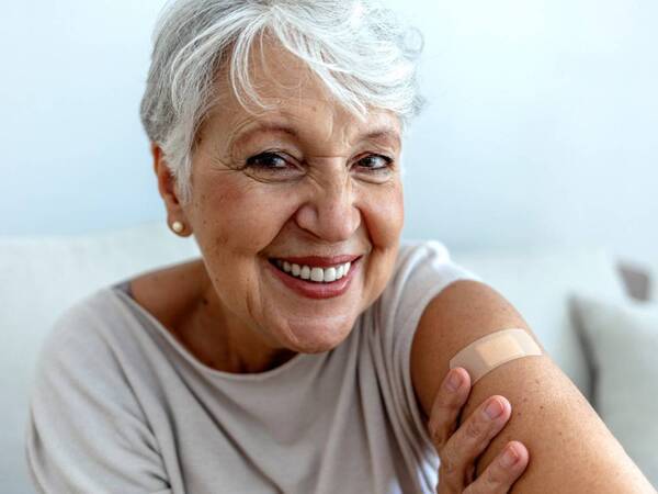 An older woman with gray hair smiles as she shows off her arm with a small bandage after her Shingles vaccine.