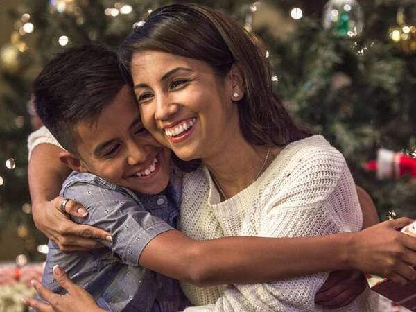 A young mother hugs her son with a Christmas tree in the background.